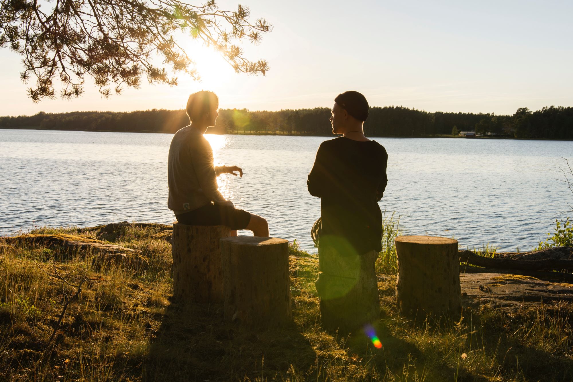 Zwei Männer sitzen am See auf Baumstümpfen und unterhalten sich. Es wirkt vertraut und friedlich.